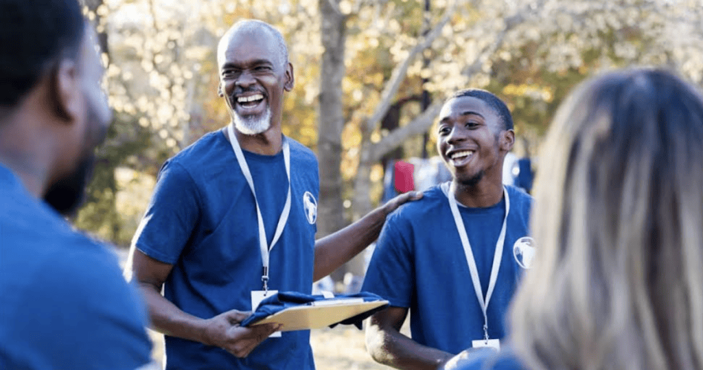 Two people wearing blue shirts and name tags smile and interact outdoors with others in the background.