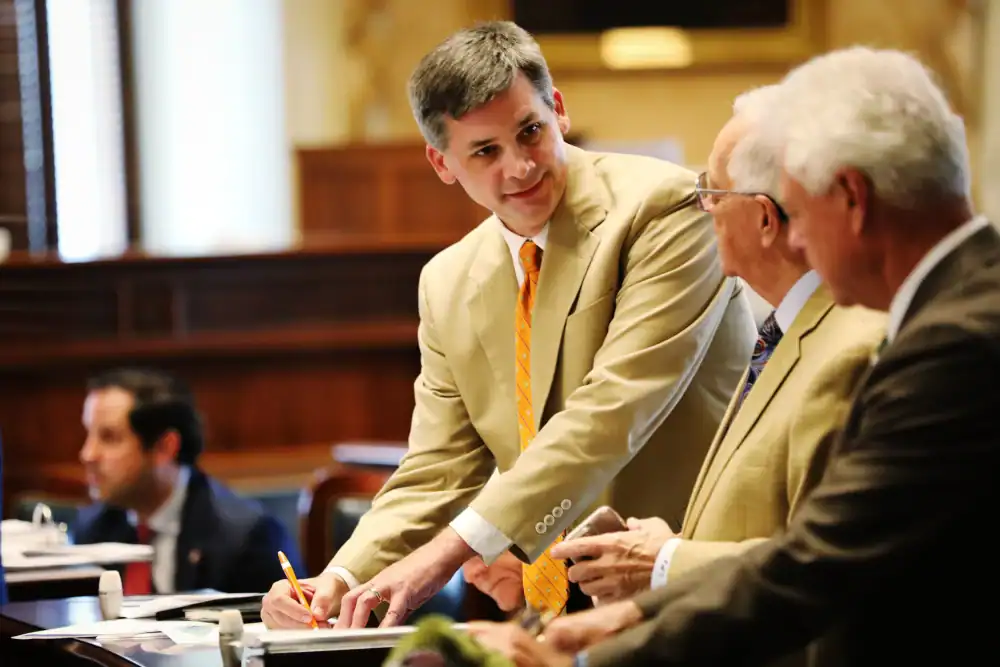 Three men in suits converse at a desk in a formal setting; one man stands and writes with a pencil while the others face him.