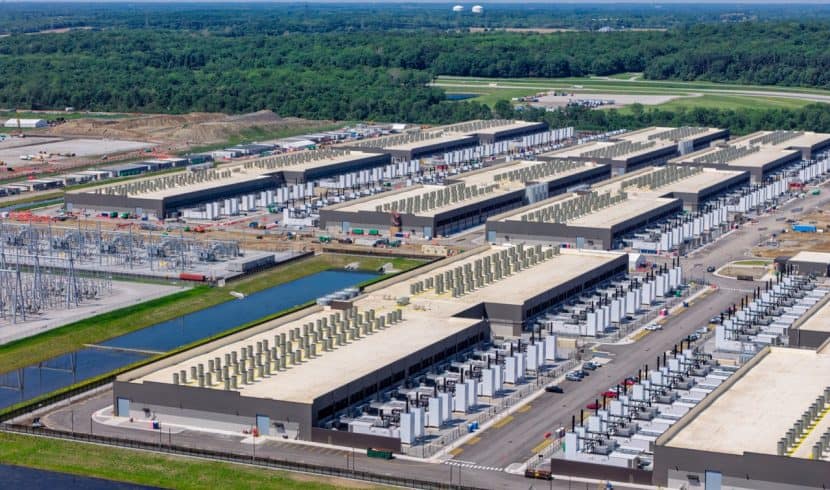 Aerial view of a large data center complex with multiple buildings, cooling units on rooftops, parking areas, and nearby power infrastructure, surrounded by greenery.