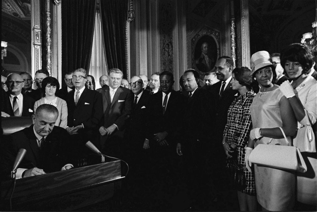 A group of men and women observe as a man seated at a desk signs a document in a formal, ornate room.